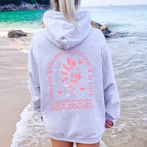 May include: Gray hooded sweatshirt with a pink graphic print on the back. The graphic features a rose with a ribbon and the text "And the greatest of these is love 1 Corinthians 13:13 Faith Hope Love".
