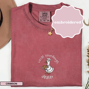 Puede incluir: Camiseta de color carmes&iacute; con un dise&ntilde;o bordado. El dise&ntilde;o presenta un pato de dibujos animados con un gorro de chef y sosteniendo un pan, con las palabras "Local sourdough dealer" en un patr&oacute;n circular alrededor del pato.
