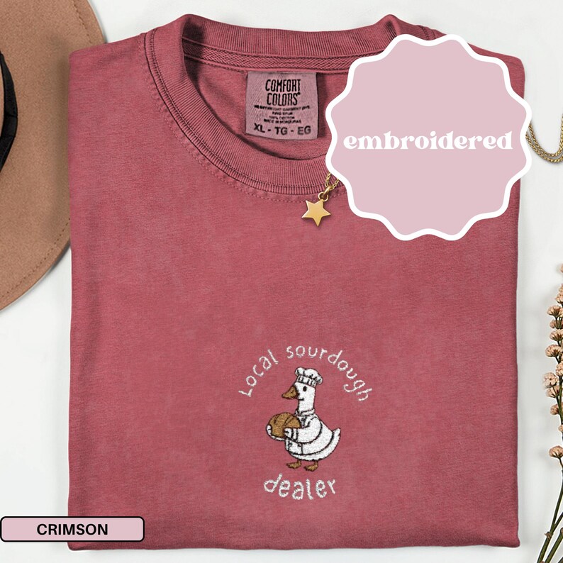 Puede incluir: Camiseta de color carmes&iacute; con un dise&ntilde;o bordado. El dise&ntilde;o presenta un pato de dibujos animados con un gorro de chef y sosteniendo un pan, con las palabras "Local sourdough dealer" en un patr&oacute;n circular alrededor del pato.