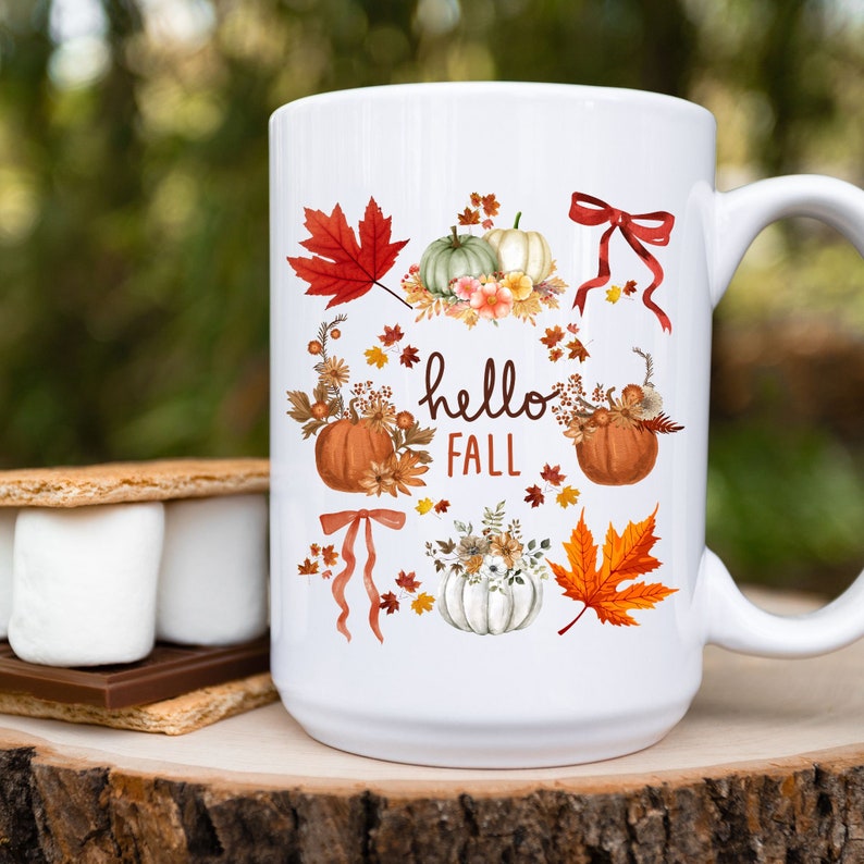 May include: White ceramic mug with a fall-themed design. The mug features pumpkins, leaves, and flowers in shades of orange, red, green, and brown. The text "hello fall" is printed on the mug.
