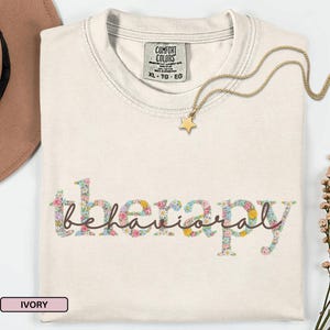 May include: Ivory-colored t-shirt with the words "behavioral therapy" in a floral design. A gold star pendant necklace rests on the shirt. The shirt has a Comfort Colors tag.