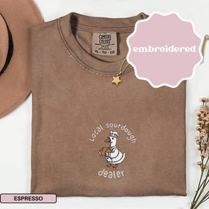 Puede incluir: Camiseta color espresso con un dise&ntilde;o bordado de un ganso con gorro de chef y pan, con las palabras "Local sourdough dealer". La camiseta tiene cuello redondo y una etiqueta que dice "Comfort Colors". Se ve un collar de estrella dorada.