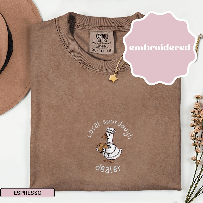 Puede incluir: Camiseta color espresso con un dise&ntilde;o bordado de un ganso con gorro de chef y pan, con las palabras "Local sourdough dealer". La camiseta tiene cuello redondo y una etiqueta que dice "Comfort Colors". Se ve un collar de estrella dorada.