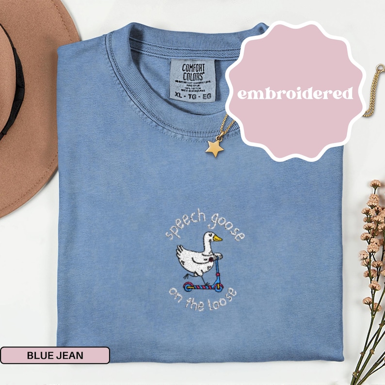 May include: A blue jean Comfort Colors t-shirt with an embroidered design. The design features a white goose riding a scooter with the text "speech goose on the loose". The shirt is size XL.