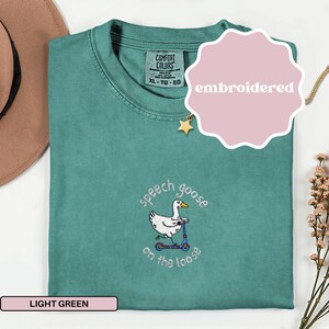May include: Light green t-shirt with an embroidered design of a goose on a scooter, with the text "speech goose on the loose". The shirt is labeled "Comfort Colors" and is size XL-TG-EG.