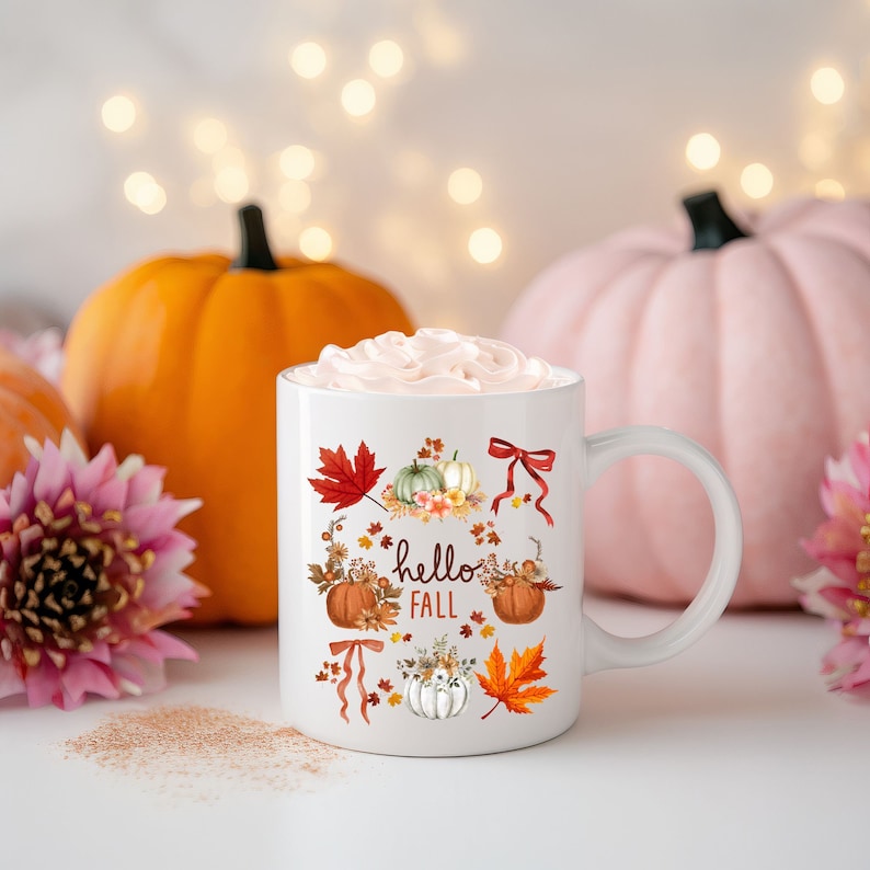 May include: White ceramic mug with a fall-themed design featuring pumpkins, leaves, and the text "hello fall". The mug is filled with a pink whipped cream topping and is surrounded by pumpkins.