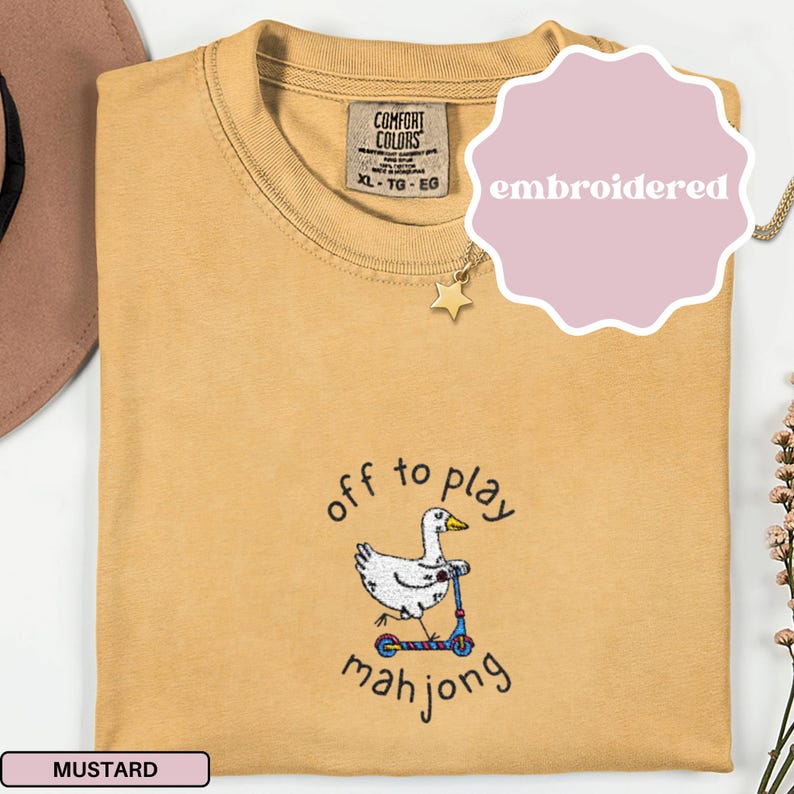 May include: Mustard-colored Comfort Colors t-shirt with an embroidered design. The design features a white duck riding a scooter, with the words "off to play mahjong" in black script. The shirt has a tag that says XL-TG-EG.