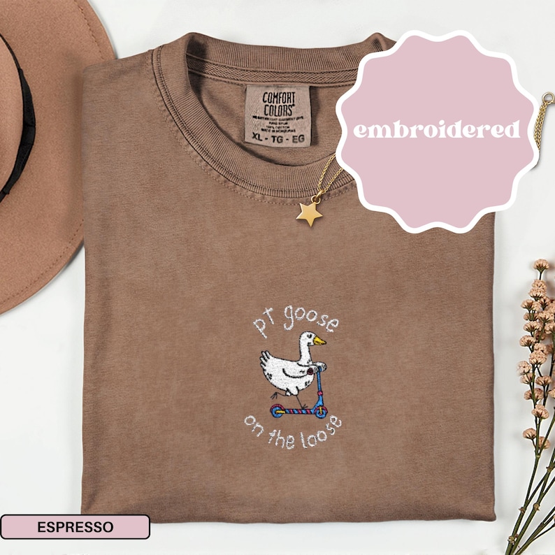 May include: Espresso-colored Comfort Colors t-shirt with an embroidered design. The design features a white goose riding a blue scooter, with the text "pt goose on the loose". A gold star necklace and a pink "embroidered" sticker are also visible.
