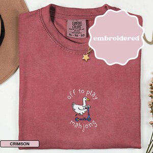 May include: Crimson-colored t-shirt with an embroidered design. The design features a white goose riding a blue scooter, with the words "off to play mahjong" in a whimsical font. The shirt has a crew neck and a tag that reads XL-TG-EG.
