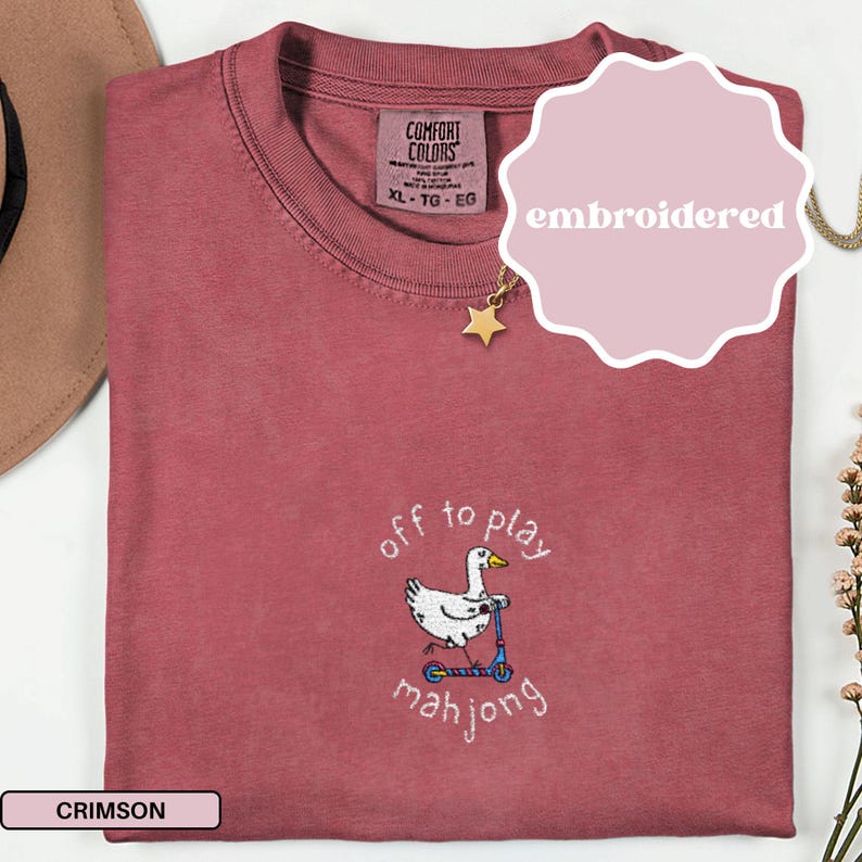 May include: Crimson-colored t-shirt with an embroidered design. The design features a white goose riding a blue scooter, with the words "off to play mahjong" in a whimsical font. The shirt has a crew neck and a tag that reads XL-TG-EG.