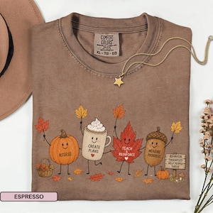 May include: A brown short-sleeve t-shirt featuring a graphic of a pumpkin, a coffee mug, a maple leaf, and an acorn with faces. The text reads ASSESS, CREATE PLANS, TEACH & REINFORCE, MEASURE PROGRESS, and BEHAVIOR THERAPISTS HELP PEOPLE THRIVE.