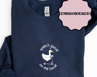 Puede incluir: Sudadera azul marino con diseño bordado. El diseño presenta un ganso blanco montado en un patinete, con las palabras "speech goose on the loose". La palabra "EMBROIDERED" está en un círculo rosa. La sudadera es azul marino.