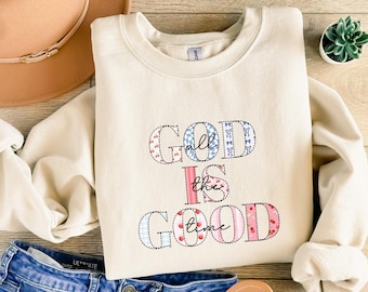 May include: A cream-coloured sweatshirt featuring the embroidered phrase "God is Good all the time" in blue, pink, and white. The image includes blue denim shorts, a brown hat, and sandals, creating a casual aesthetic.