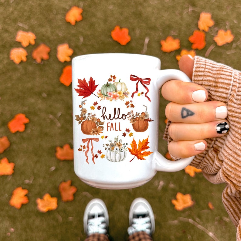 May include: White ceramic coffee mug with a fall-themed design. The mug features a watercolor illustration of pumpkins, leaves, and the text "hello fall".