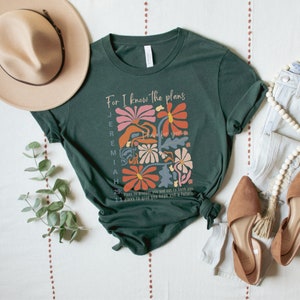 May include: A green t-shirt with a floral design and the text "For I know the plans I have for you" Jeremiah 29:11. The text is in a white font and the flowers are in shades of pink, orange, and blue.