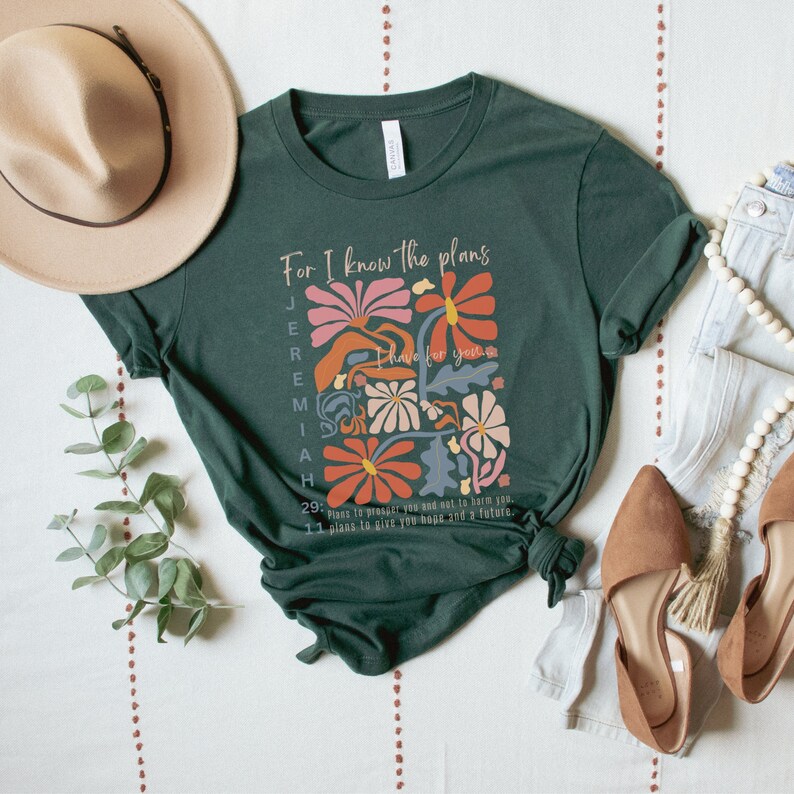May include: A green t-shirt with a floral design and the text "For I know the plans I have for you" Jeremiah 29:11. The text is in a white font and the flowers are in shades of pink, orange, and blue.