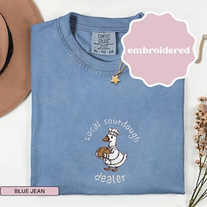 May include: A blue jean Comfort Colors t-shirt with an embroidered design. The design features a cartoon duck wearing a chef's hat and holding a loaf of bread, with the words "Local sourdough dealer". The shirt has a crew neck and short sleeves.