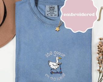 May include: A blue jean Comfort Colors t-shirt with an embroidered design. The design features a white goose riding a scooter with the text "aba goose on the loose". The shirt has a crew neck and a tag that says XL-TG-EG.