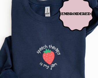 Puede incluir: Sudadera azul marino bordada con un diseño de fresa. La fresa tiene un tallo verde y el texto "speech therapy is my jam" en blanco. La palabra "EMBROIDERED" está en un círculo rosa. La palabra "NAVY" está en un rectángulo azul marino.