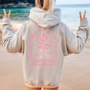 May include: A light gray hoodie with a pink graphic print of a rose with a bow and the text "And the greatest of these is love 1 Corinthians 13:13 Faith Hope Love".