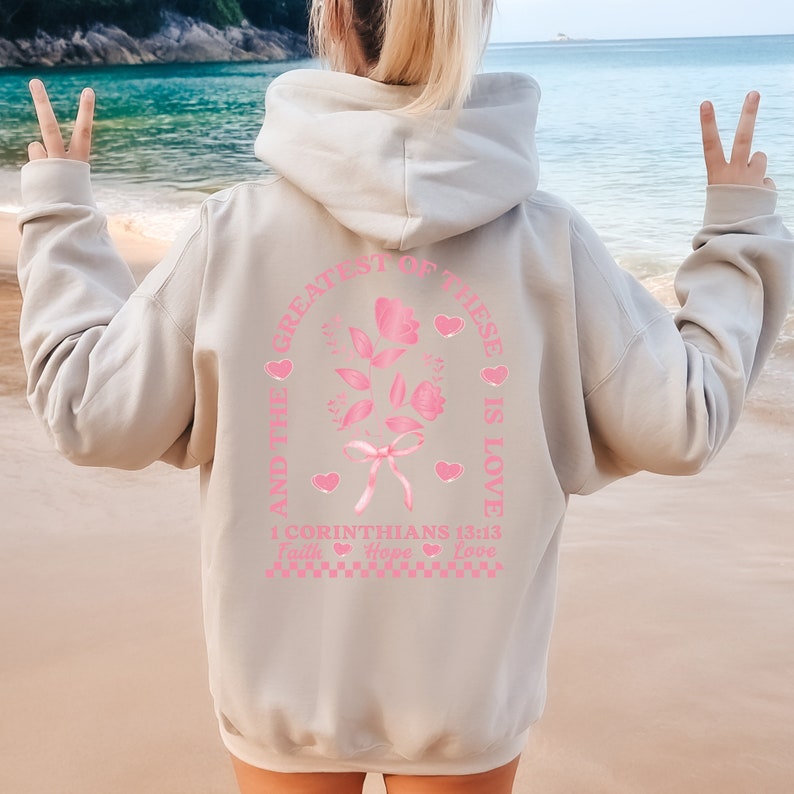May include: A light gray hoodie with a pink graphic print of a rose with a bow and the text "And the greatest of these is love 1 Corinthians 13:13 Faith Hope Love".