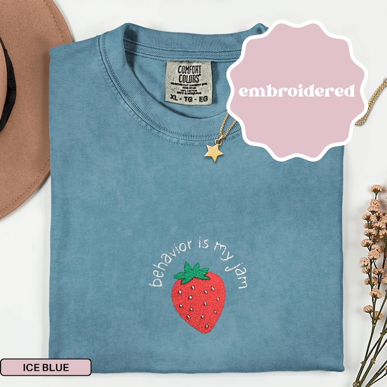 Peut inclure: T-shirt bleu glacier avec une fraise brod&eacute;e et le texte "behavior is my jam". Le t-shirt a un col rond et des manches courtes. Un collier pendentif &eacute;toile dor&eacute;e est visible. Le t-shirt est &eacute;tiquet&eacute; "XL - TG - EG".