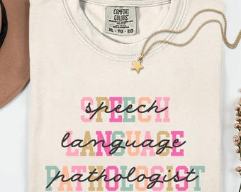 Könnte beinhalten: Elfenbeinfarbenes T-Shirt mit den Worten "SPEECH LANGUAGE PATHOLOGIST" in bunten Blockbuchstaben. Eine goldene Stern-Anhänger-Halskette liegt auf dem Shirt. Das Shirt ist sauber gefaltet.