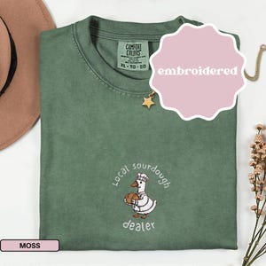 Puede incluir: Camiseta verde musgo con un dise&ntilde;o bordado que presenta un ganso de dibujos animados sosteniendo un pan, las palabras "Local sourdough dealer" y la palabra "embroidered". La camiseta tiene cuello redondo y mangas cortas.
