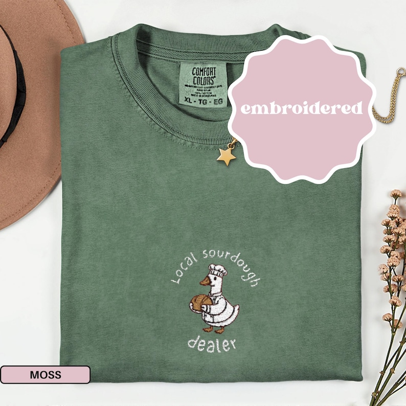Puede incluir: Camiseta verde musgo con un dise&ntilde;o bordado que presenta un ganso de dibujos animados sosteniendo un pan, las palabras "Local sourdough dealer" y la palabra "embroidered". La camiseta tiene cuello redondo y mangas cortas.