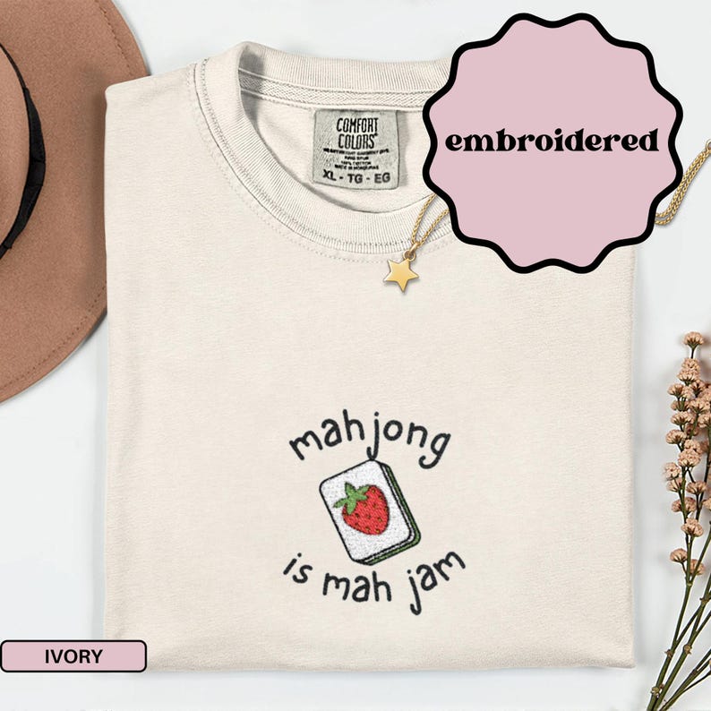 May include: Ivory-colored t-shirt with the text "mahjong is mah jam" and an embroidered strawberry on a mahjong tile. The shirt has a Comfort Colors tag and a gold star necklace.