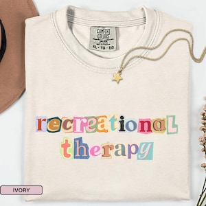 以下が含まれることがあります： アイボリー色のTシャツ。カラフルな切り抜き文字のデザインで「recreational therapy」の文字がプリントされています。金の星のペンダントネックレスがTシャツの上に置かれています。TシャツにはComfort Colorsのタグが付いており、きれいに折りたたまれています。