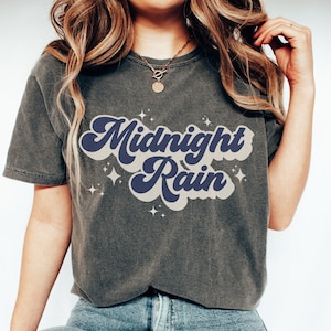 May include: Dark grey t-shirt featuring the text "Midnight Rain" in a retro font, with navy blue and light grey colours. The design includes small star graphics. A casual wear item.