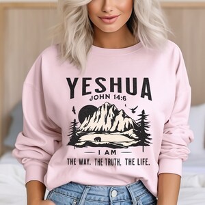 May include: Pink sweatshirt with a black graphic print of a mountain range, a bear, and birds. The text "YESHUA JOHN 14:6 I AM THE WAY. THE TRUTH. THE LIFE." is printed on the sweatshirt.