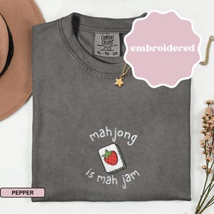 May include: Pepper-colored Comfort Colors t-shirt with an embroidered design. The design features a mahjong tile with a red strawberry and the text "mahjong is mah jam". The shirt has a crew neck and short sleeves. A gold necklace with a star pendant is visible.