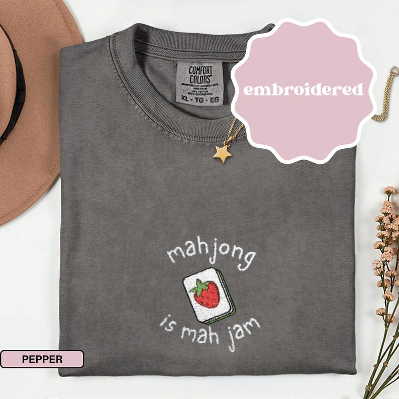 May include: Pepper-colored Comfort Colors t-shirt with an embroidered design. The design features a mahjong tile with a red strawberry and the text "mahjong is mah jam". The shirt has a crew neck and short sleeves. A gold necklace with a star pendant is visible.