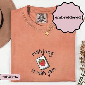 May include: Terracotta-colored, embroidered t-shirt with the text "mahjong is mah jam" and a graphic of a mahjong tile with a strawberry. The shirt has a crew neck and is labeled XL-TG-EG.