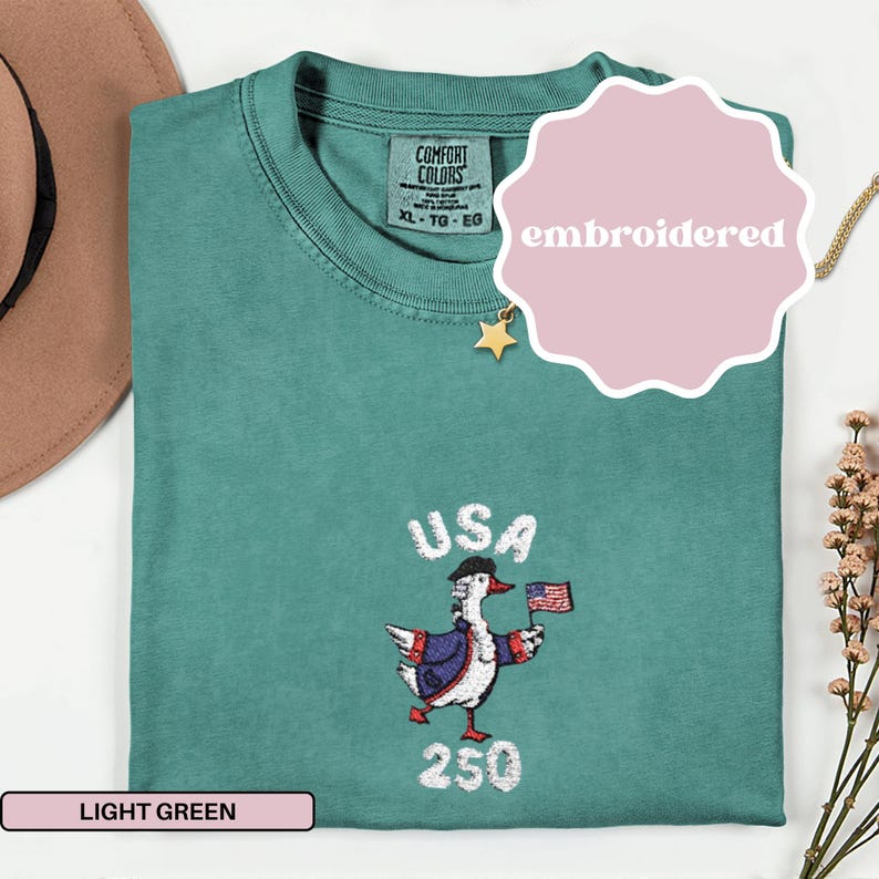 May include: Light green Comfort Colors t-shirt with an embroidered design. The design features a goose wearing a hat and jacket, holding a small American flag, with the text "USA 250". The shirt is labeled "XL - TG - EG".