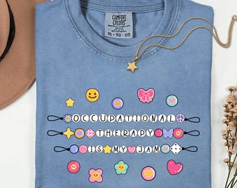 Op de afbeelding: Een blauw spijkerbroekkleurig t-shirt met de woorden "OCCUPATIONAL THERAPY IS MY JAM" in witte letters. Het shirt heeft kleurrijke grafische ontwerpen, waaronder een smiley, harten en bloemen. Een gouden ketting met een sterhanger is over het shirt gedrapeerd.