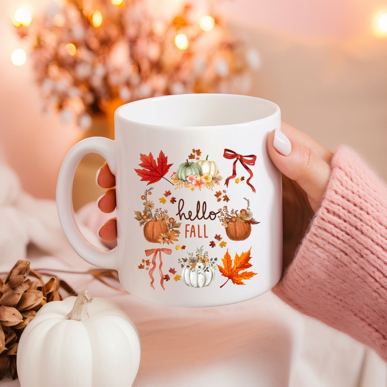 May include: White ceramic coffee mug with a fall-themed design. The mug features pumpkins, leaves, and the text "hello fall".