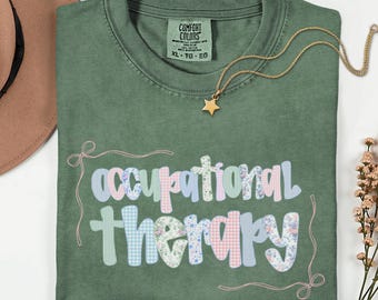 Op de afbeelding: Een mosgroen t-shirt met de woorden "occupational therapy" in een kleurrijk, patroonontwerp. Het shirt heeft een ronde hals en korte mouwen. Een gouden ketting met een sterhanger en een bruine hoed zijn ook zichtbaar.