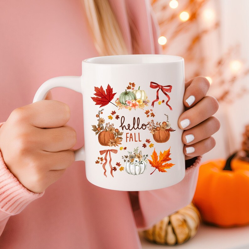 May include: White ceramic mug with a fall-themed design. The mug features pumpkins, leaves, and flowers in shades of orange, brown, and green. The text "hello fall" is printed on the mug.