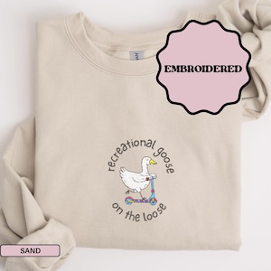 May include: A sand-colored crewneck sweatshirt featuring an embroidered graphic of a white goose riding a blue and pink scooter. The text "recreational goose on the loose" is embroidered in a circular pattern around the goose illustration.