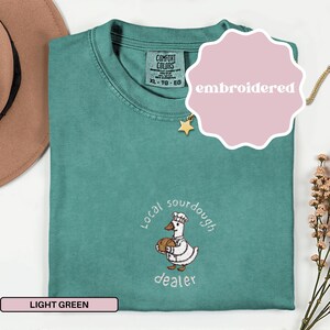 Puede incluir: Camiseta verde claro con un dise&ntilde;o bordado. El dise&ntilde;o presenta un ganso de dibujos animados con gorro de chef y sosteniendo un pan, con las palabras "local sourdough dealer". La camiseta tambi&eacute;n tiene una etiqueta que dice "Comfort Colors".