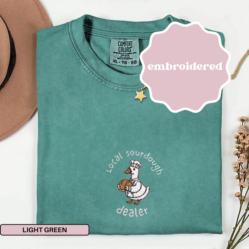 Puede incluir: Camiseta verde claro con un dise&ntilde;o bordado. El dise&ntilde;o presenta un ganso de dibujos animados con gorro de chef y sosteniendo un pan, con las palabras "local sourdough dealer". La camiseta tambi&eacute;n tiene una etiqueta que dice "Comfort Colors".