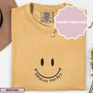 Può includere: Una t-shirt a girocollo color giallo senape con una faccina sorridente ricamata in nero e la scritta "behavior therapy" sottostante. La maglia presenta un'etichetta del marchio Comfort Colors sul colletto interno e un piccolo ciondolo a stella dorata.