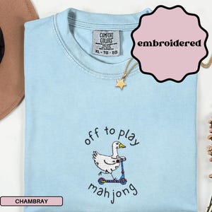 May include: Light blue chambray t-shirt with the text "off to play mahjong" and an embroidered graphic of a goose on a scooter. The shirt has a crew neck and short sleeves. A gold star pendant necklace is also visible.