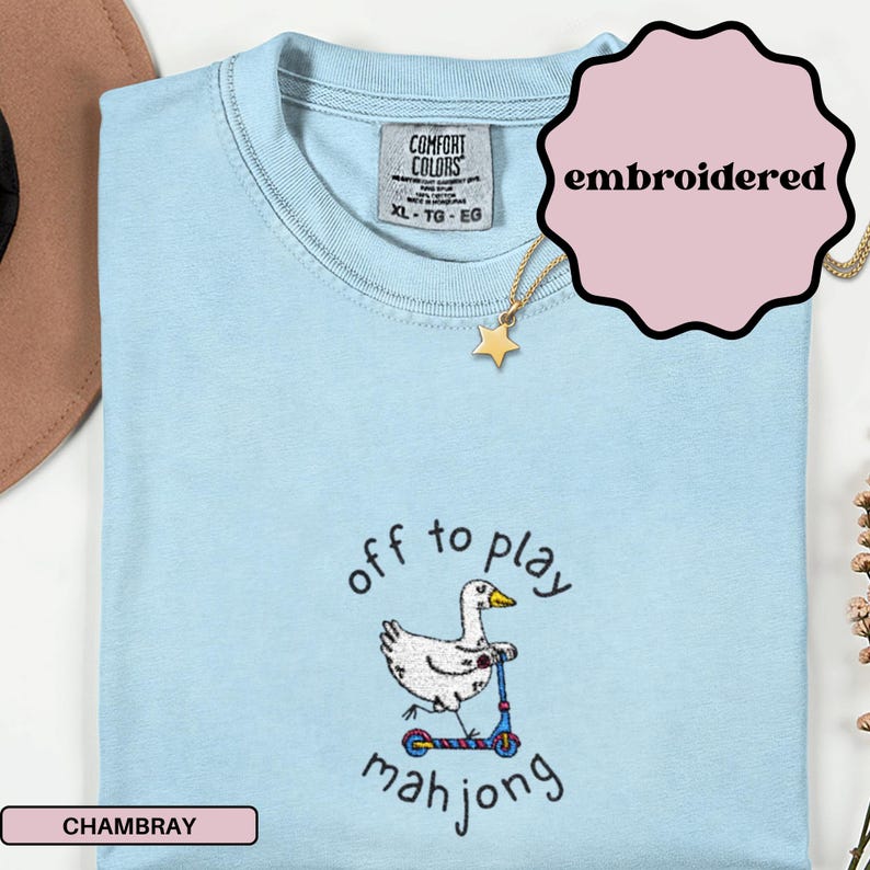 May include: Light blue chambray t-shirt with the text "off to play mahjong" and an embroidered graphic of a goose on a scooter. The shirt has a crew neck and short sleeves. A gold star pendant necklace is also visible.