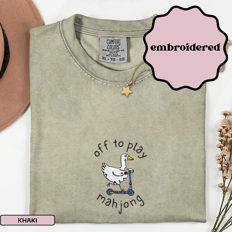 May include: Khaki-colored, embroidered t-shirt with the text "off to play mahjong" and a cartoon duck riding a scooter. The shirt has a Comfort Colors tag and a gold star necklace. The word "embroidered" is in a pink bubble.