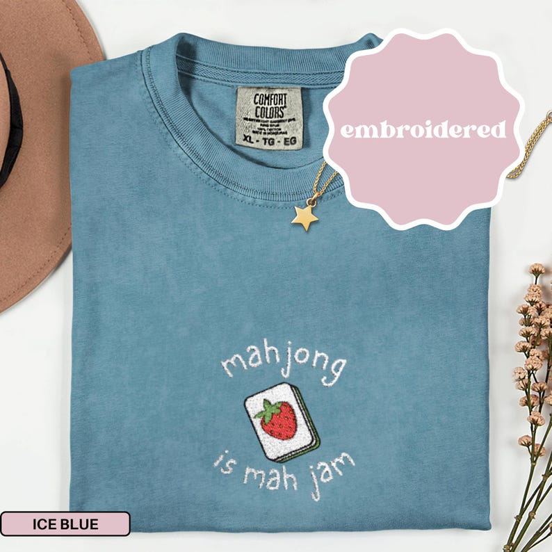 May include: Ice blue Comfort Colors t-shirt with embroidered text and a mahjong tile graphic. The text reads "mah jong is mah jam". A gold star necklace is visible. The shirt is size XL-TG-EG.