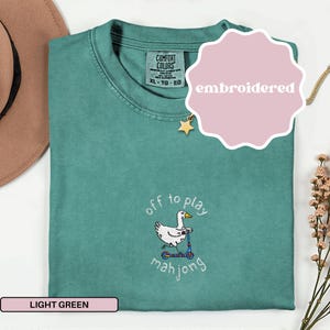 May include: Light green Comfort Colors t-shirt with an embroidered design. The design features a white duck riding a blue scooter with the words "off to play mahjong". The shirt has a tag that says XL-TG-EG.